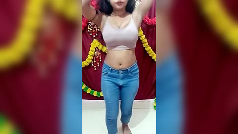 Media: Video of a woman with medium skin tone and shoulder-length black hair, wearing a white crop top and blue jeans, standing in front of a festive red backdrop adorned with yellow garlands and greenery, with blurred figures in red and yellow attire in the foreground.