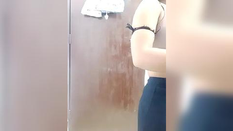 Media: Video of a partially blurred, fair-skinned woman with a slim physique in a sleeveless white top and dark pants, standing in a dimly lit, metallic bathroom with a towel hanging on a hook.