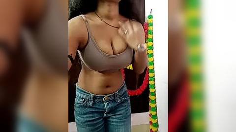 Media: Video of a curvaceous woman with medium-dark skin, long black hair, wearing a beige crop top and blue jeans, holding a flower garland. Background includes a colorful floral arrangement and blurred figures.