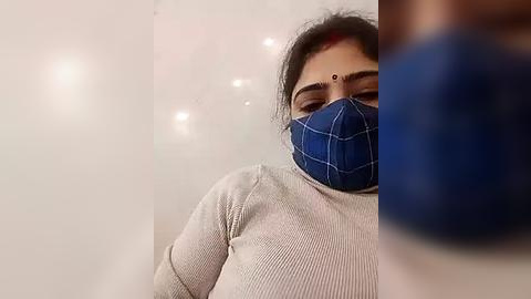 Media: Video of a woman with medium skin tone, wearing a blue plaid mask, a white long-sleeve top, and a red bindi, standing indoors with a blurred background.