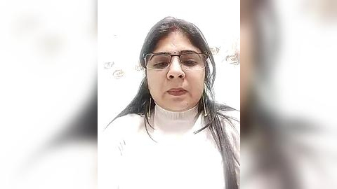 Media: A video of a middle-aged South Asian woman with long black hair, wearing rectangular glasses and a white turtleneck sweater, against a blurred background.