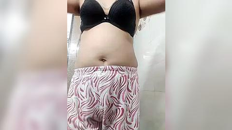 Media: Video of a woman in a black bra, showing her midriff, and wearing patterned pajama pants, standing in a bathroom with a tiled wall and a white shower curtain.