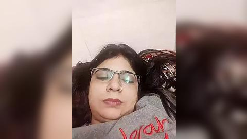 Media: Video of a woman with long black hair, wearing glasses, lying on a bed with a gray blanket, blurry background.