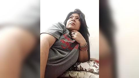 Media: Video of a woman with long black hair, wearing a gray \"MOUNTAIN\" t-shirt, sitting on a bed with patterned sheets, looking contemplative.