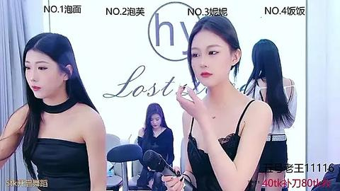 Media: Video of two Asian women with long black hair, applying makeup. One wears a black strapless dress, the other a black top. Background includes a white wall with Chinese text and a mirror.