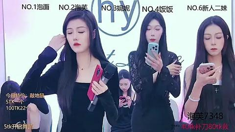 Media: Video of four young East Asian women, all with long black hair, holding smartphones, wearing black dresses, in a brightly lit room with white walls.