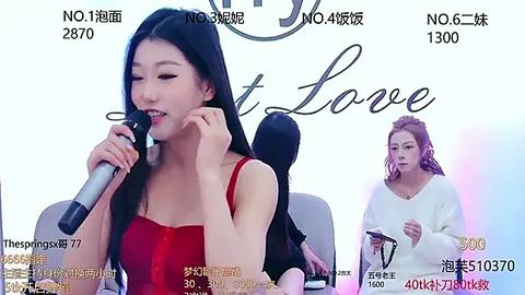 Media: Video of a fair-skinned East Asian woman with long black hair, wearing a red dress, speaking into a microphone at a press conference. Background includes another woman in a white sweater and blurred audience members.