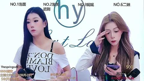 Media: Video of two East Asian women with long, straight hair, one in a white off-shoulder top, the other in a white jacket, both looking down at their smartphones.