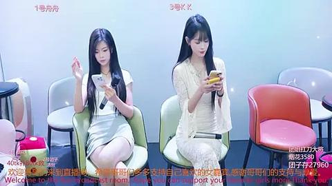 Media: Video of two young East Asian women with long black hair, seated in modern chairs, using smartphones, in a minimalist room with pastel-colored chairs.