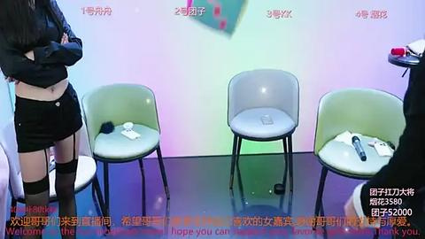 Media: Video of an exhibition room with three green chairs, a person in black attire, and a white phone on a table, under a colorful light background.