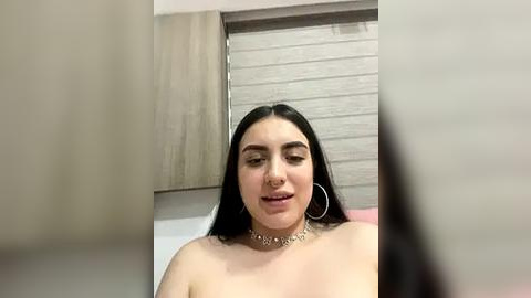 Media: Video of a young woman with long black hair, wearing a silver choker and large hoop earrings, sitting topless in a room with beige walls and a window with closed blinds.