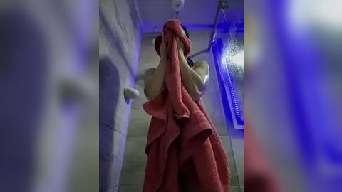 Media: A video of a woman in a shower, covering her breasts with a red towel, surrounded by blue lighting and tiled walls.