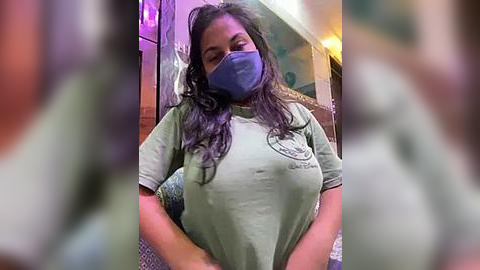 Media: Video of a young woman with long, wavy brown hair, wearing a light green t-shirt, a blue face mask, and blue gloves, standing indoors with blurred background, wearing a blue face mask.