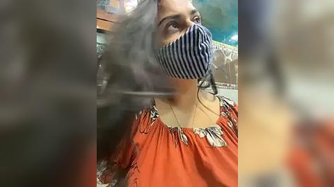 Media: Video of a woman with medium skin tone wearing a black and white striped face mask, a red blouse with floral embroidery, and a necklace. Background blurred, possibly indoors.