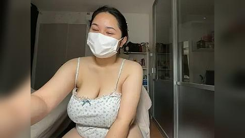 Media: Video of an Asian woman with medium skin tone, wearing a white face mask, floral camisole, and sitting in a bedroom with a white bed, glass wardrobe, and shelves.