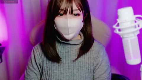 Media: A video of a young Asian woman with long, straight brown hair, wearing a gray ribbed sweater and a white mask, seated indoors with a purple and pink background, and a white microphone on her left.