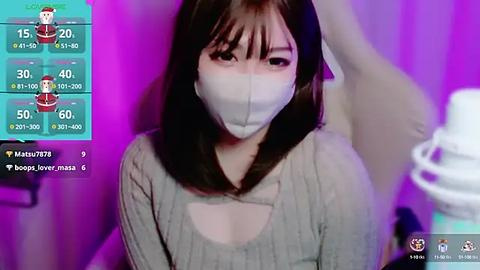 Media: Video of an East Asian woman with shoulder-length black hair, wearing a gray ribbed sweater and a white surgical mask, standing indoors with a purple wall background.