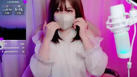 Media: A video of an East Asian woman with long, straight brown hair and fair skin, wearing a white mask, white blouse, and white pearl necklace. Background includes a pink wall and a white microphone stand.