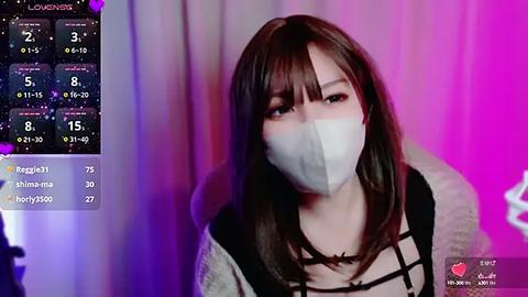 Media: A video of a young Asian woman with long brown hair, wearing a face mask, black top, and grey cardigan, against a pink and purple background.