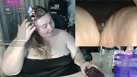 Media: Video of a plus-sized woman with fair skin, long blonde hair, wearing a black strapless top, smiling while using a phone, in a messy room.