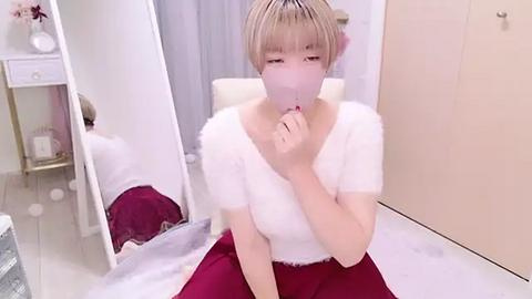 Media: Video of a young Asian woman with fair skin, short blonde hair, wearing a white fluffy top and red skirt, holding a mask to her mouth, sitting on a white chair in a minimalist, white-walled room.