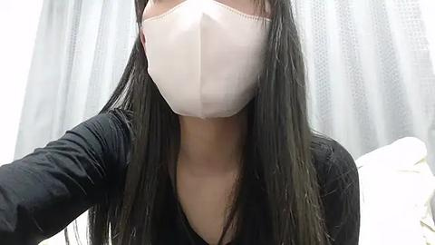 Media: Video of a woman with long, dark hair, wearing a white surgical mask and a black top, standing indoors with light-colored curtains in the background.