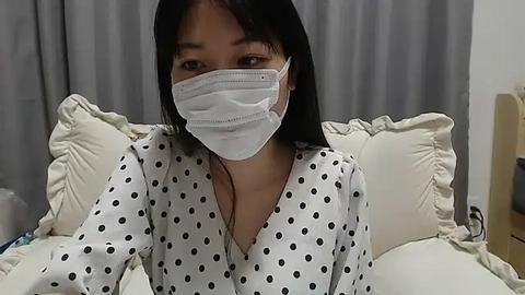 Media: Video of a young Asian woman with long black hair, wearing a white polka-dot nightgown and a white surgical mask, lying in a hospital bed with white pillows and gray curtains in the background.