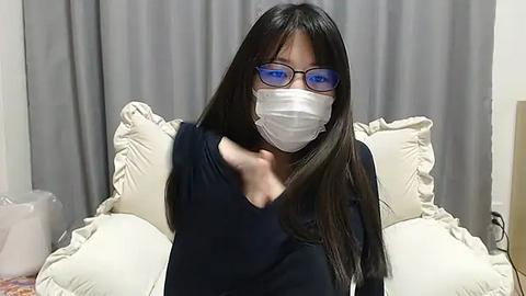 Media: Video of an Asian woman with long black hair, blue-framed glasses, and a white face mask, wearing a black top, sitting on a bed with white pillows and gray curtains in the background.