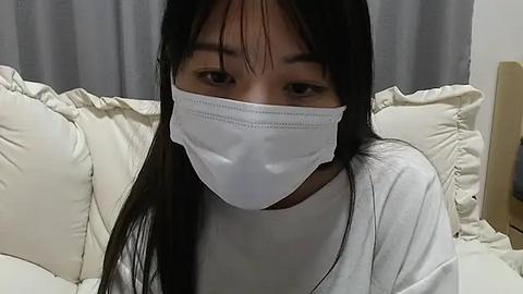 Media: A video of an Asian woman with long black hair and a white face mask, wearing a white T-shirt, sitting on a white bed with gray curtains in the background.