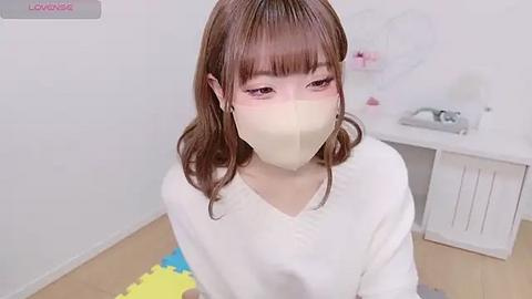 Media: A video of a young Asian woman with light skin, brown hair, and a surgical mask, sitting in a clean, white room with a sink and blue mat on the floor.