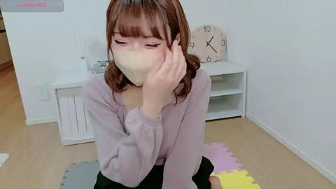 Media: Video of a young Asian woman with shoulder-length brown hair, wearing a lavender sweater, sitting on a colorful rug, blowing her nose with a tissue in a minimalist, white-walled room.