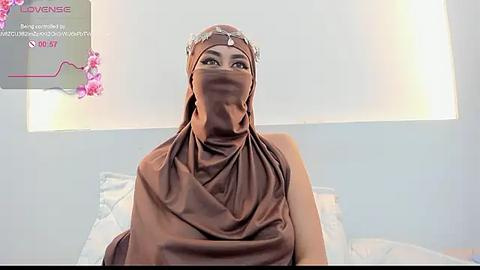 Media: A video of a woman with light skin wearing a brown hijab and veil, sitting on a bed with white sheets in a bright, minimalistic room.