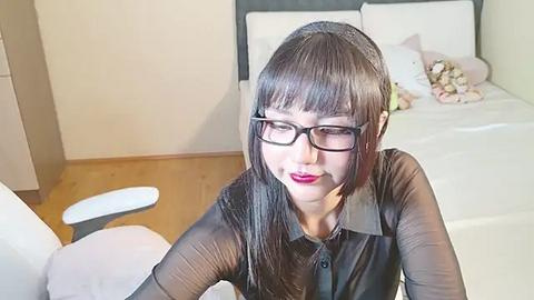 Media: Video of an Asian woman with straight black hair, wearing black glasses, a black blouse, and red lipstick, sitting on a white chair in a minimalist bedroom with a neatly made bed.