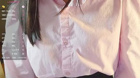 Media: Video of a woman's chest, wearing a light pink blouse with visible buttons and collar. Her dark hair partially covers her shoulder. Background includes a blurred window with sunlight streaming in.