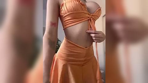 Media: Video of a slender, light-skinned woman in a low-cut, orange halter top and matching skirt, standing indoors. Her breasts are moderately sized, and her hand rests on her hip. The background is blurred.