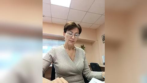 Media: A video of a middle-aged woman with short brown hair, wearing glasses and a light gray ribbed sweater, sitting at a desk in a bright office with fluorescent lighting and beige walls.