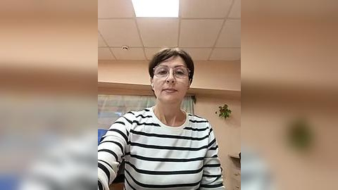 Media: Video of a middle-aged woman with short brown hair, wearing glasses and a black-and-white striped sweater, standing in an office with fluorescent lights, beige walls, and a potted plant in the background.