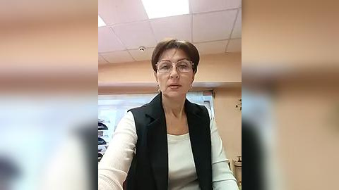 Media: Video of a middle-aged woman with short brown hair, wearing glasses, a black vest over a white shirt, and a serious expression, in an office with beige walls, fluorescent lighting, and a blurred background.