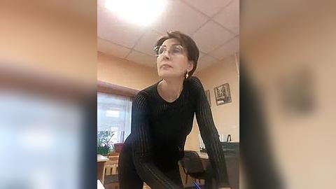 Media: Video of a woman with short dark hair, wearing a black ribbed sweater, leaning forward in a cluttered, beige room with a tiled ceiling and a window with blinds.