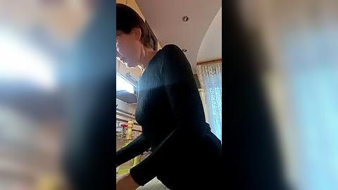 Media: A video of a woman in a black long-sleeve shirt, leaning over a countertop, blurred by a camera lens. The background shows a kitchen with cabinets and a window with sheer curtains.