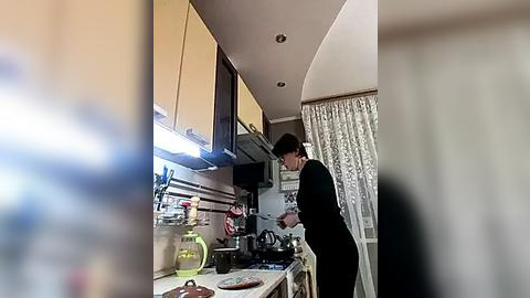 Media: A video of a woman in a kitchen with a blurred background. She is wearing black clothing and is cooking. The kitchen has modern cabinets, a sink, and a stove.