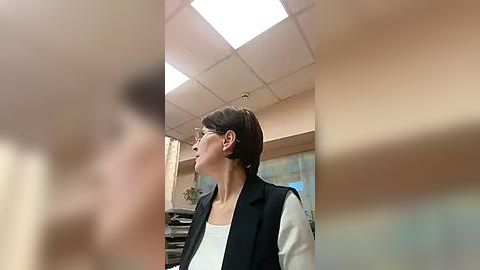 Media: A video of a woman with short dark hair, glasses, and a black blazer over a white shirt, standing in a beige office with fluorescent lights, blurred background.