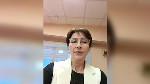 Media: Video of a middle-aged woman with short brown hair, glasses, and wearing a white blazer over a black top, standing in a room with a beige ceiling and a window with light blue curtains in the background.