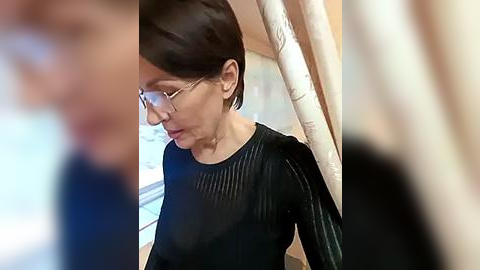 Media: Video of a woman with short dark hair, glasses, wearing a black, long-sleeved top, seated indoors, blurred background.