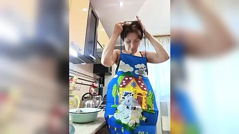 Media: Video of a woman in a bright blue apron with a colorful, cartoonish house design, adjusting her hair in a modern kitchen with stainless steel appliances and white walls.