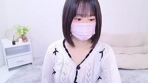 Media: Video of an East Asian woman with straight black hair, pale skin, and a pink surgical mask, wearing a white, patterned dress. Background includes a white nightstand with a vase and a beige couch.