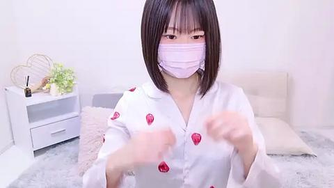 Media: Video of an Asian woman with straight black hair, wearing a pink surgical mask, white shirt with red strawberries, and gloves, in a minimalistic, light-colored room.