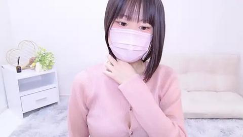 Media: Video of an East Asian woman with straight black hair and bangs, wearing a pink face mask and cardigan, standing in a minimalist room with a white wall, a white shelf, and a beige sofa.