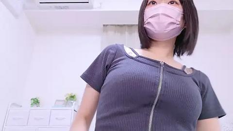 Media: Video of an East Asian woman with shoulder-length black hair, wearing a pink surgical mask and gray ribbed top, standing in a modern, bright room with white walls, air conditioner, and white dresser with green plants.