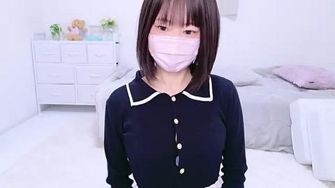 Media: A video of an Asian woman with auburn bob haircut and light skin, wearing a navy blue button-up shirt, pink face mask, and white gloves, standing in a white-walled bedroom with a dresser and teddy bear.